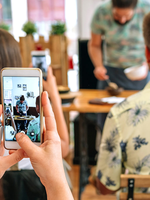 Woman taking photo with mobile to the cook in a cooking workshop. Selective focus on mobile in the foreground